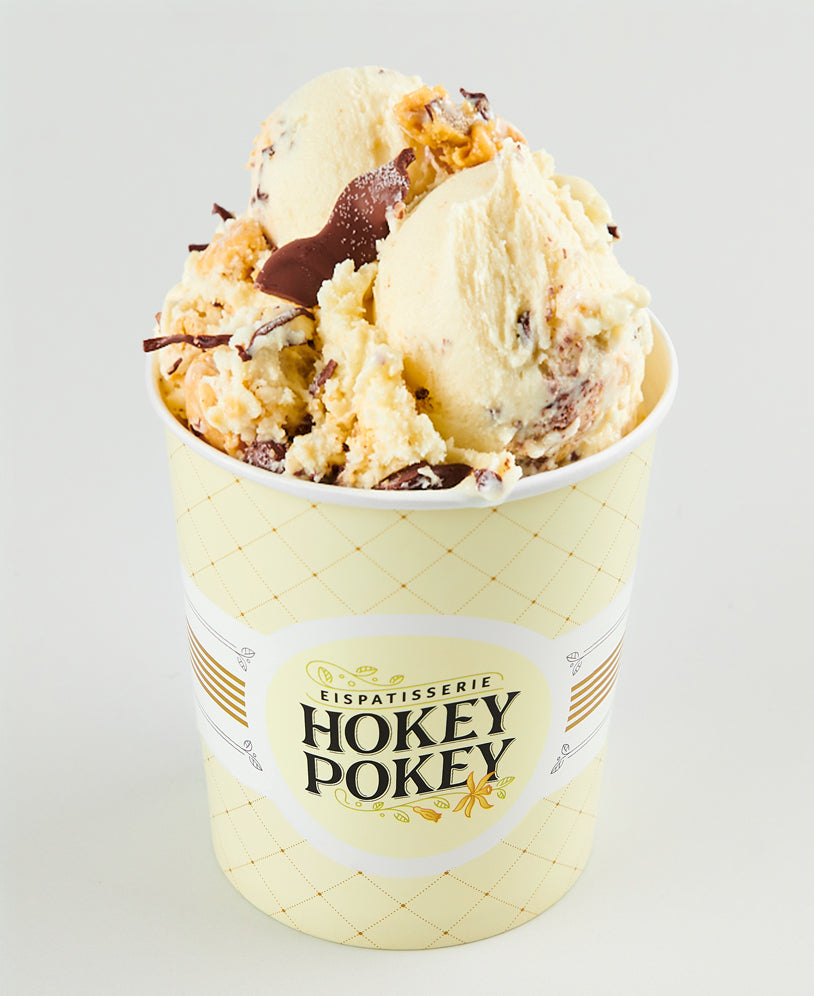 Becher mit Bananen-Erdnuss-Eis der Eispatisserie Hokey Pokey Berlin, verfeinert mit Schokostückchen und knusprigen Toppings, serviert im gelben Logo-Becher vor hellem Hintergrund.
