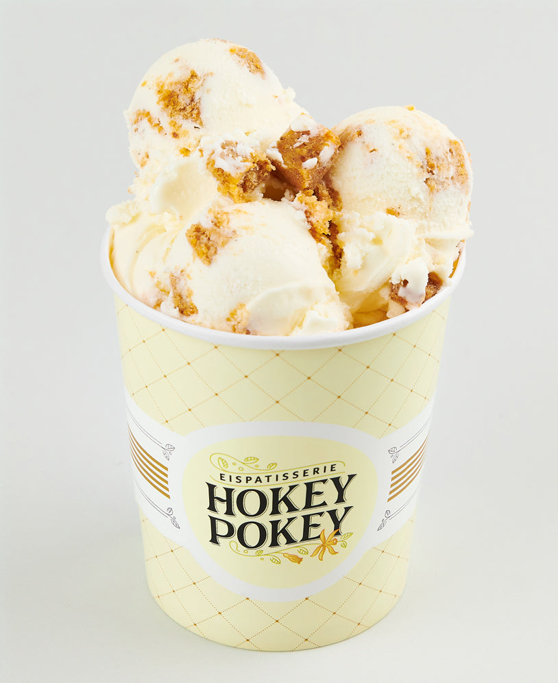 Becher mit Carrot-Cake-Eis der Eispatisserie Hokey Pokey Berlin, verfeinert mit saftigen Kuchenstücken, serviert im gelben Markenbecher vor hellem Hintergrund.