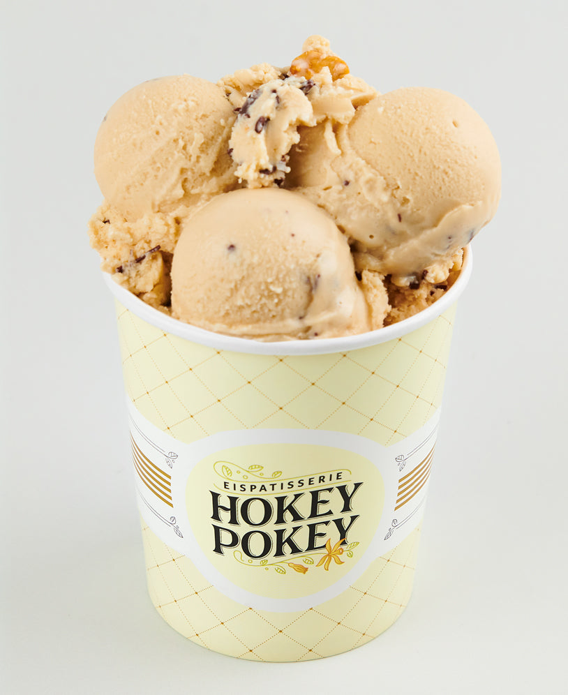 Becher mit Crunchy-Peanut-Eiscreme der Eispatisserie Hokey Pokey Berlin, verfeinert mit knackigen Erdnussstückchen und Schokosplittern, serviert im gelben Markenbecher vor hellem Hintergrund.