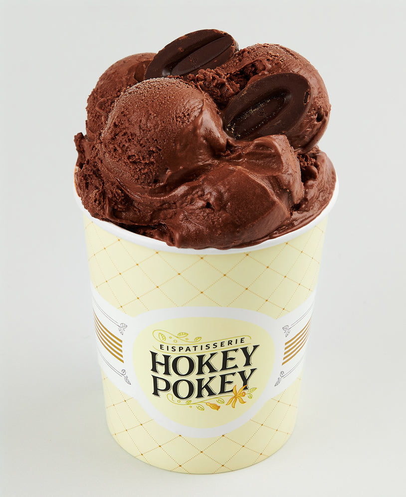 Becher mit intensiver dunkler Schokoladeneiscreme der Eispatisserie Hokey Pokey Berlin, garniert mit Schokodrops und serviert im markanten Logo-Becher auf hellem Hintergrund.