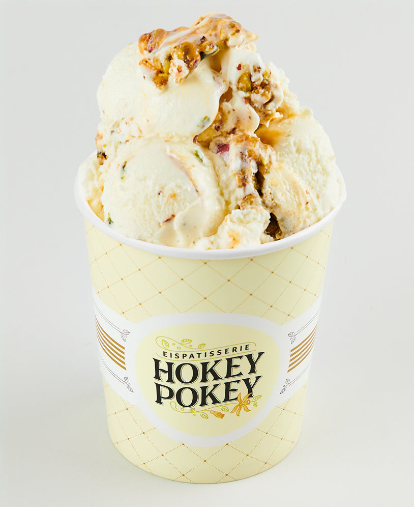 Becher mit weißer Schokoladen-Pistazieneiscreme der Eispatisserie Hokey Pokey Berlin, durchzogen mit Pistazienstückchen und Karamell, serviert im gelben Markenbecher vor hellem Hintergrund.